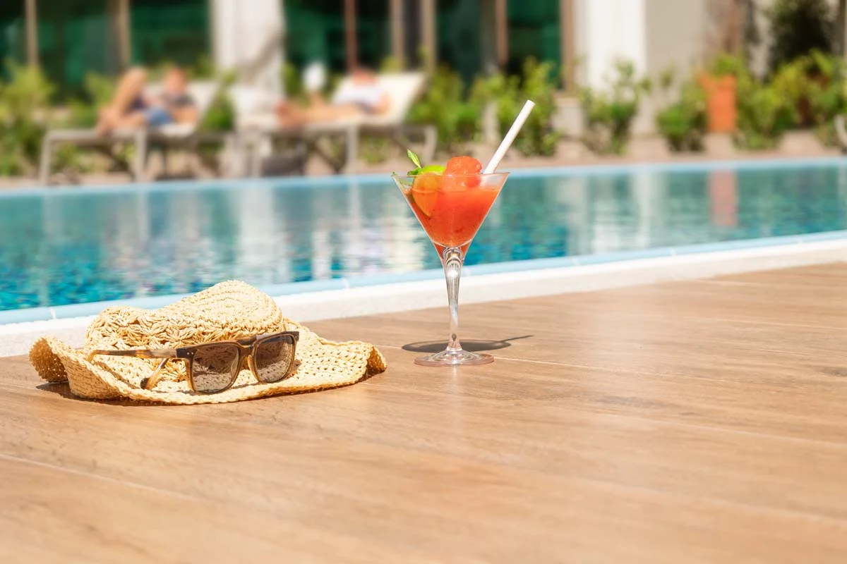 Close-up of vacation accessories and a cold drink by a bright blue swimming pool in a tropical setting