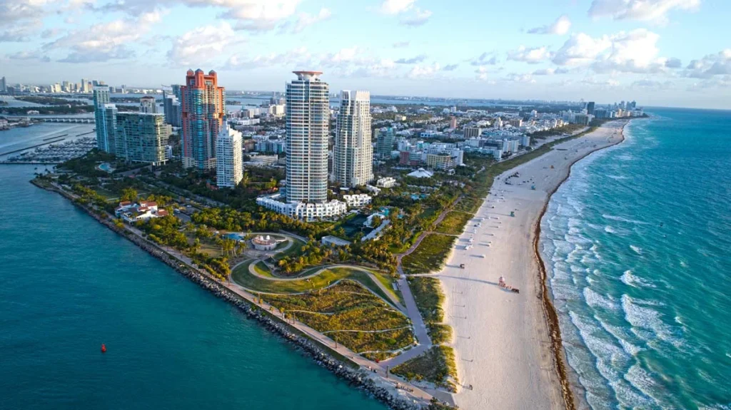 Wide aerial view of a sunny South Florida beach with luxury high-rise condos and blue ocean water