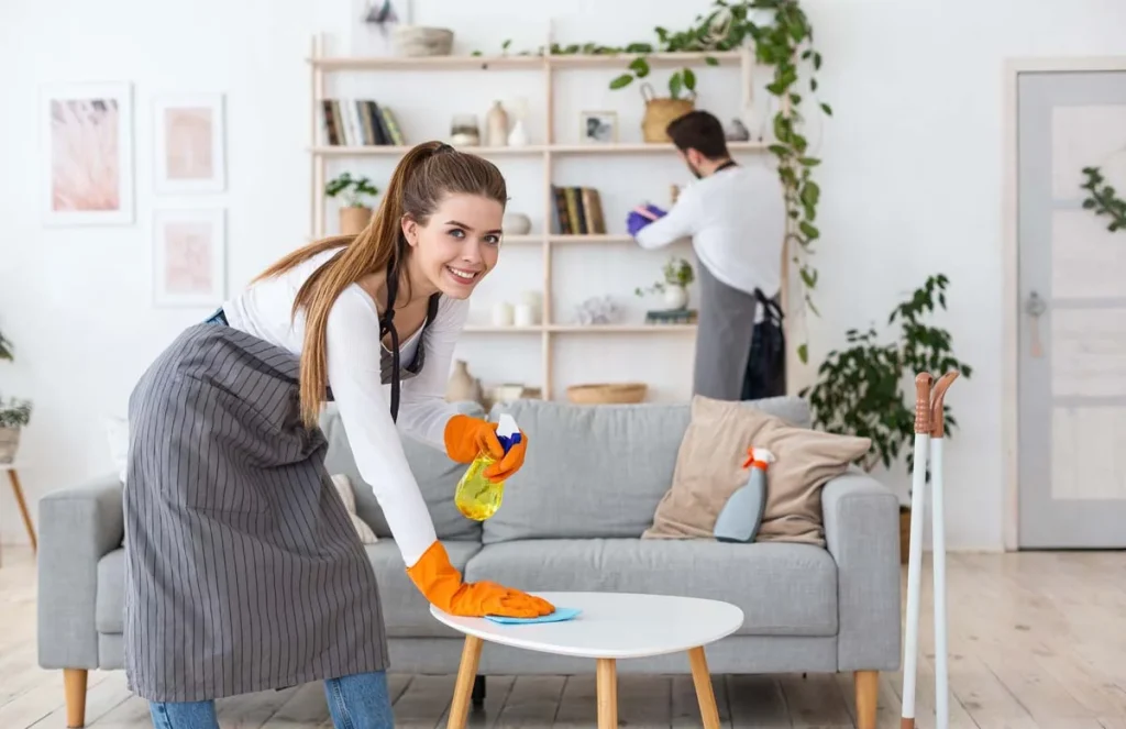 Smiling young woman in rubber gloves wipe table with a spray, a man wipes dust with a brush in the interior of a living room