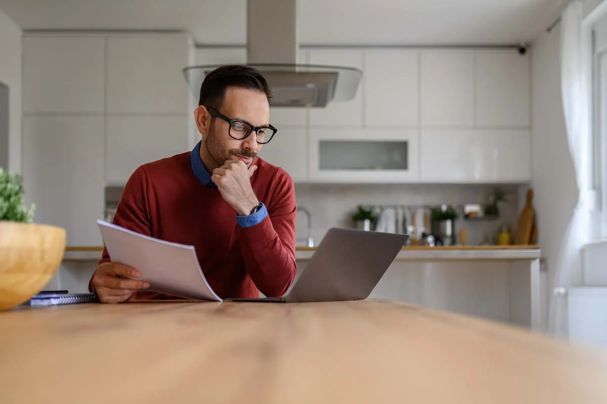 Person typing on a laptop at a kitchen counter reviewing paperwork for an online application