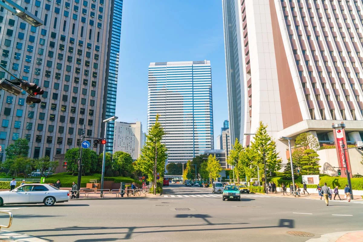 A city street lined with tall glass and stone skyscrapers, with cars, pedestrians, and trees along the sidewalk under a partly cloudy sky