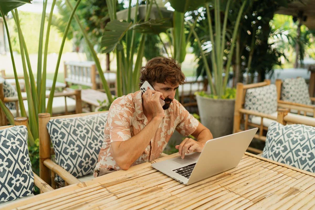 A man wearing a white shirt, types on a laptop at a wooden table, with lush green plants and a pool visible in the background