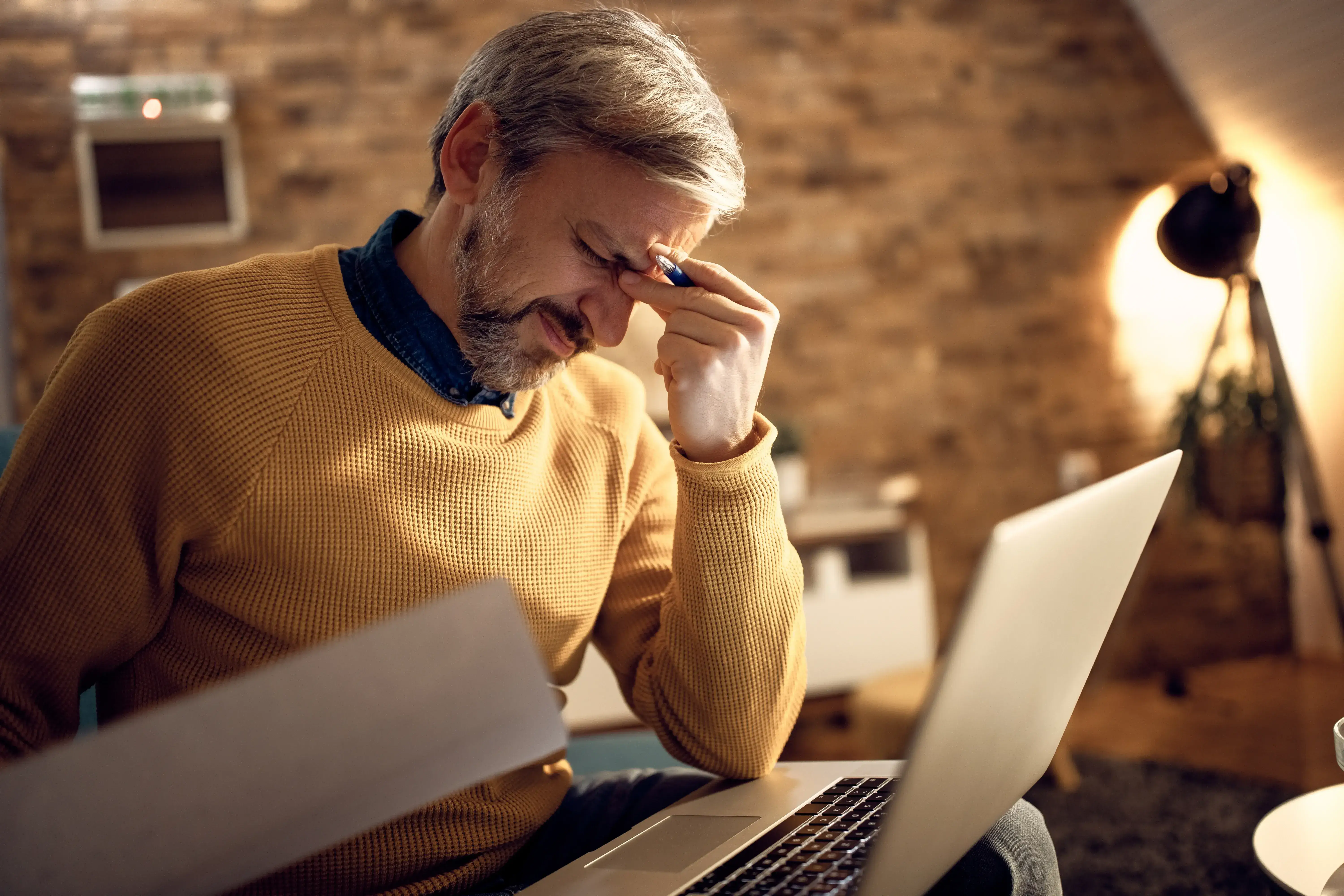 A stressed man looking over financial documents at home with a laptop on the table.