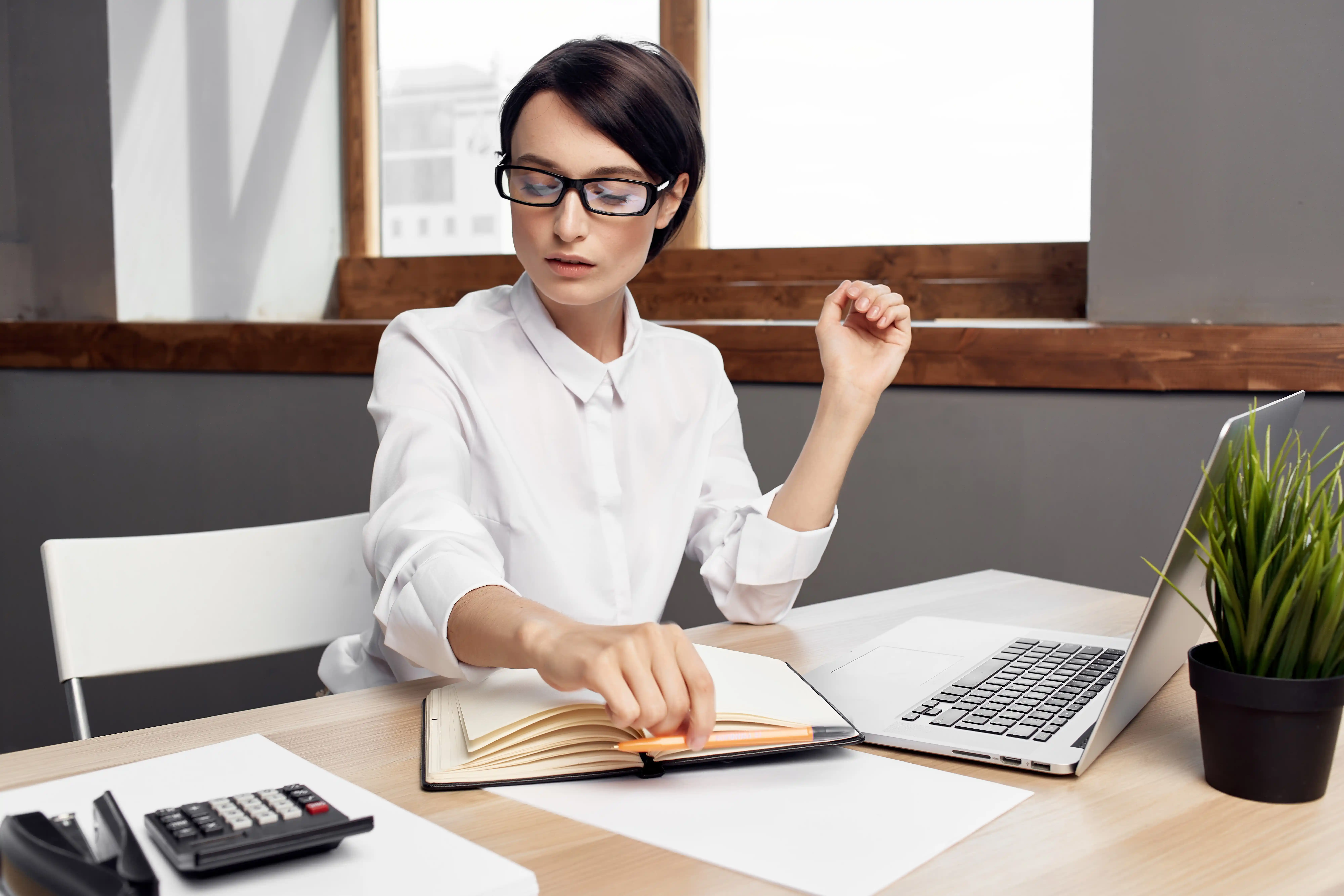 A woman with glasses writes in a notebook while using a calculator at a desk with paperwork and a laptop.