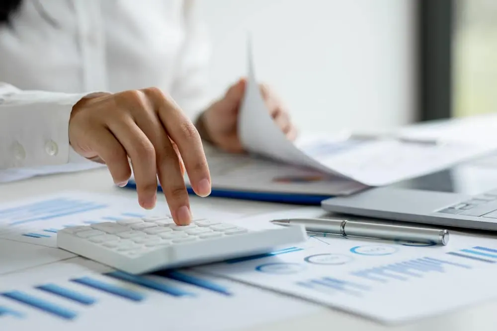 Close-up of a person in a white shirt using a calculator while reviewing financial charts and graphs on a desk, with a pen and laptop nearby