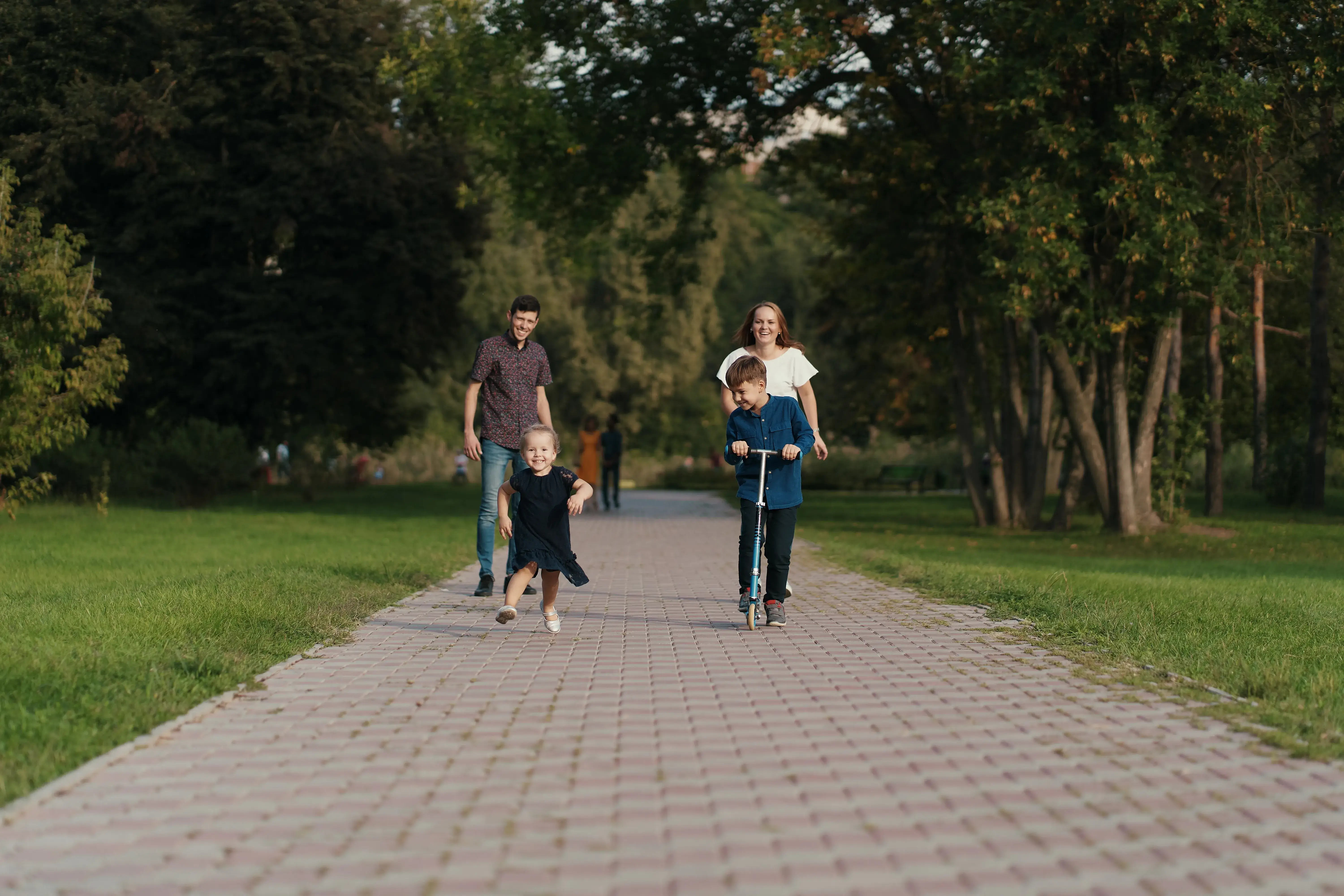 Family walking down a tree-lined residential sidewalk, with a boy riding a scooter in front