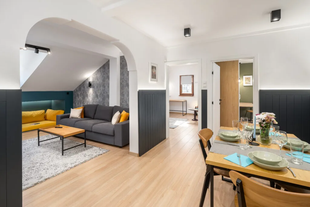 Modern open-plan living and dining space with a charcoal sectional sofa, mustard accent pillows, and light wood flooring, leading to a set dining table
