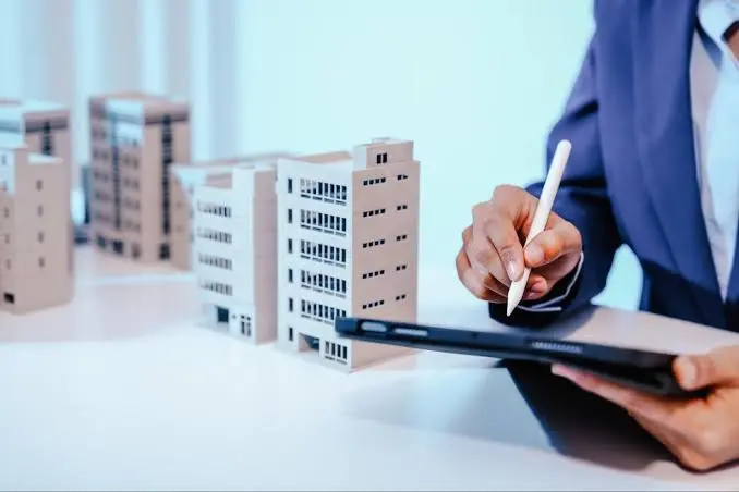 Person in a business suit using a stylus on a tablet beside a row of miniature building models, representing urban planning or real estate development