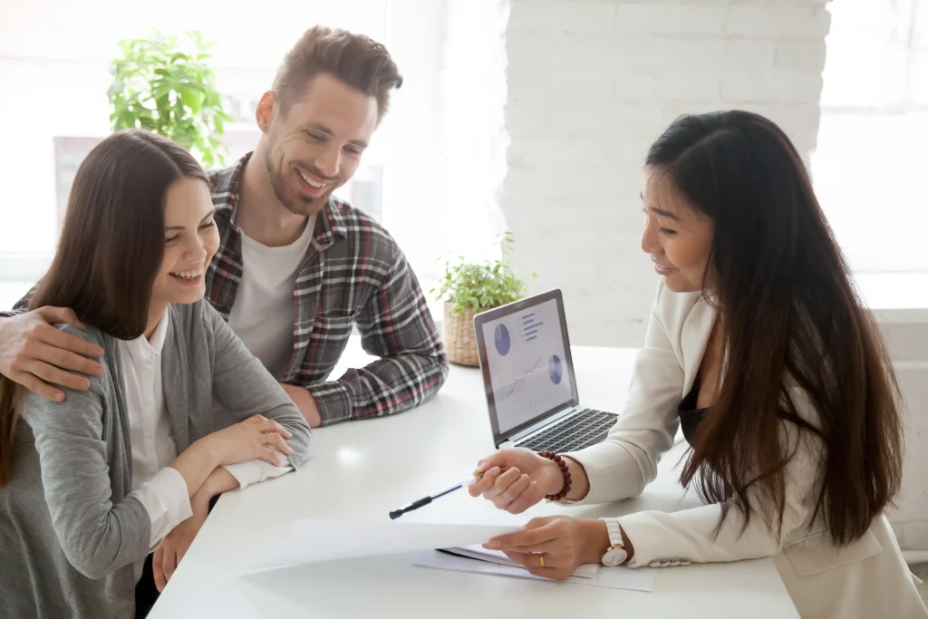 A female consultant or agent reviews paperwork with a young couple at a desk with a laptop.