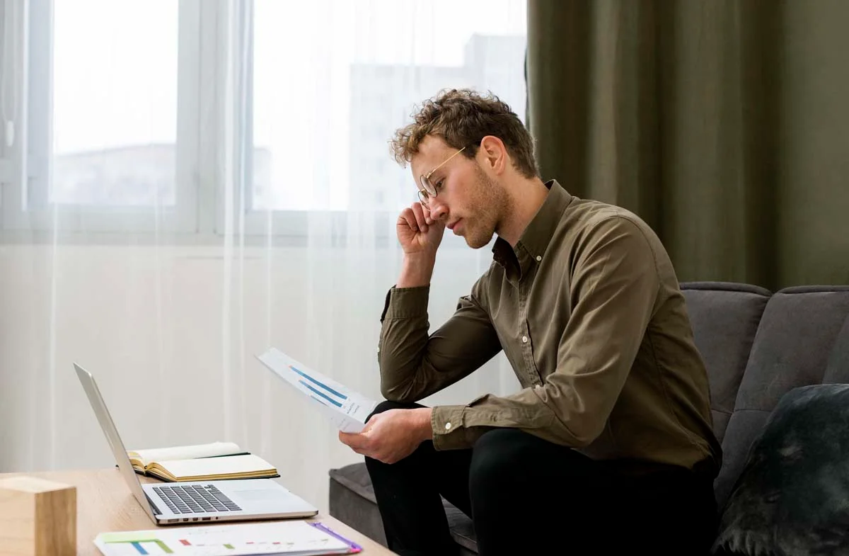 A man looking stressed while reviewing financial papers at a table next to a laptop