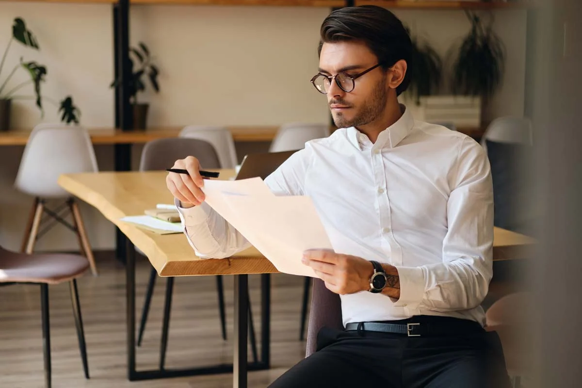 A man sits at a wooden desk, focused on reviewing a paper document in his hands