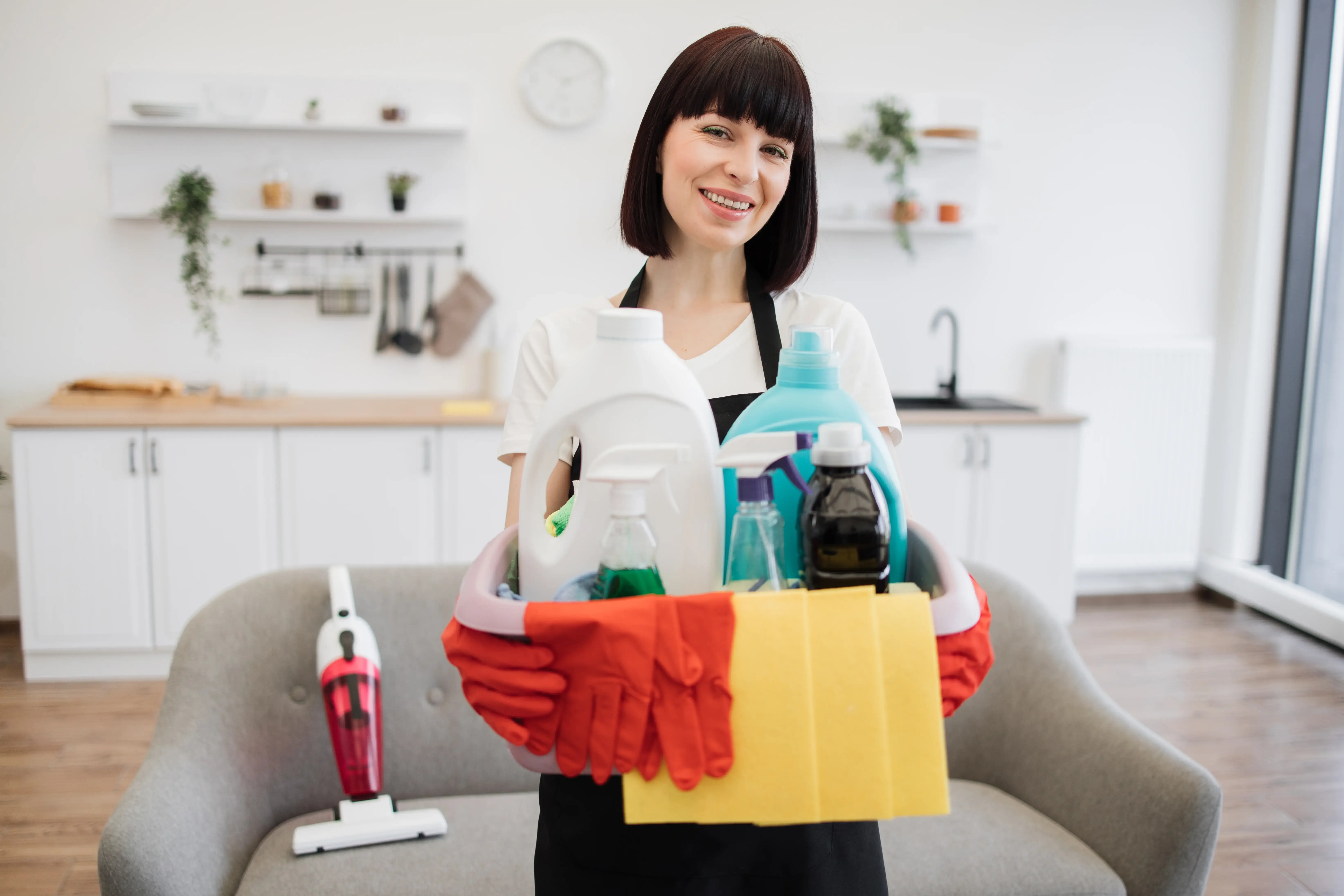 A person wearing cleaning gloves holding a bucket packed with various cleaning products and supplies.