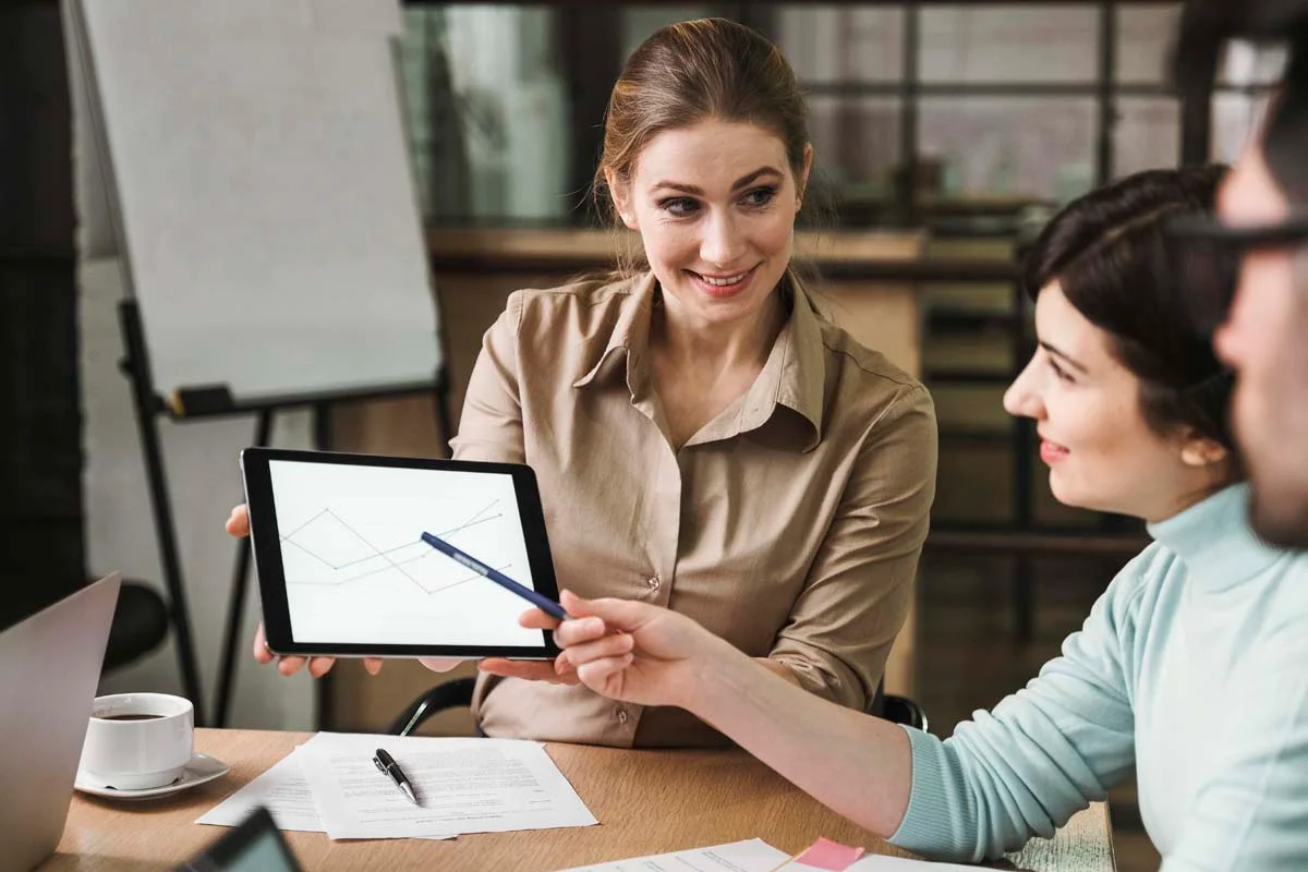 A professional woman points at a tablet screen during a meeting with a smiling young couple in a bright office