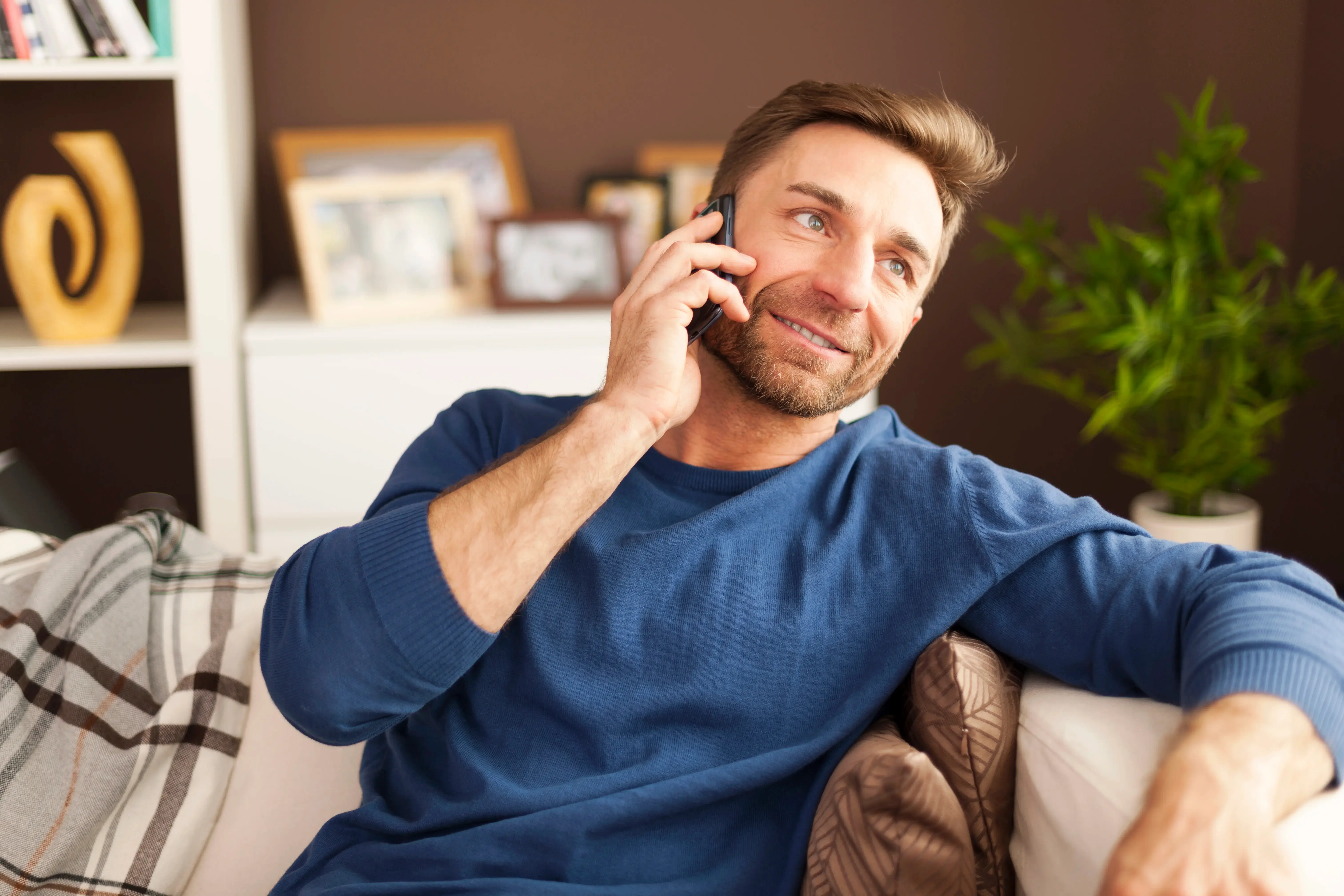 A smiling man talking on his smartphone while sitting on a living room sofa.
