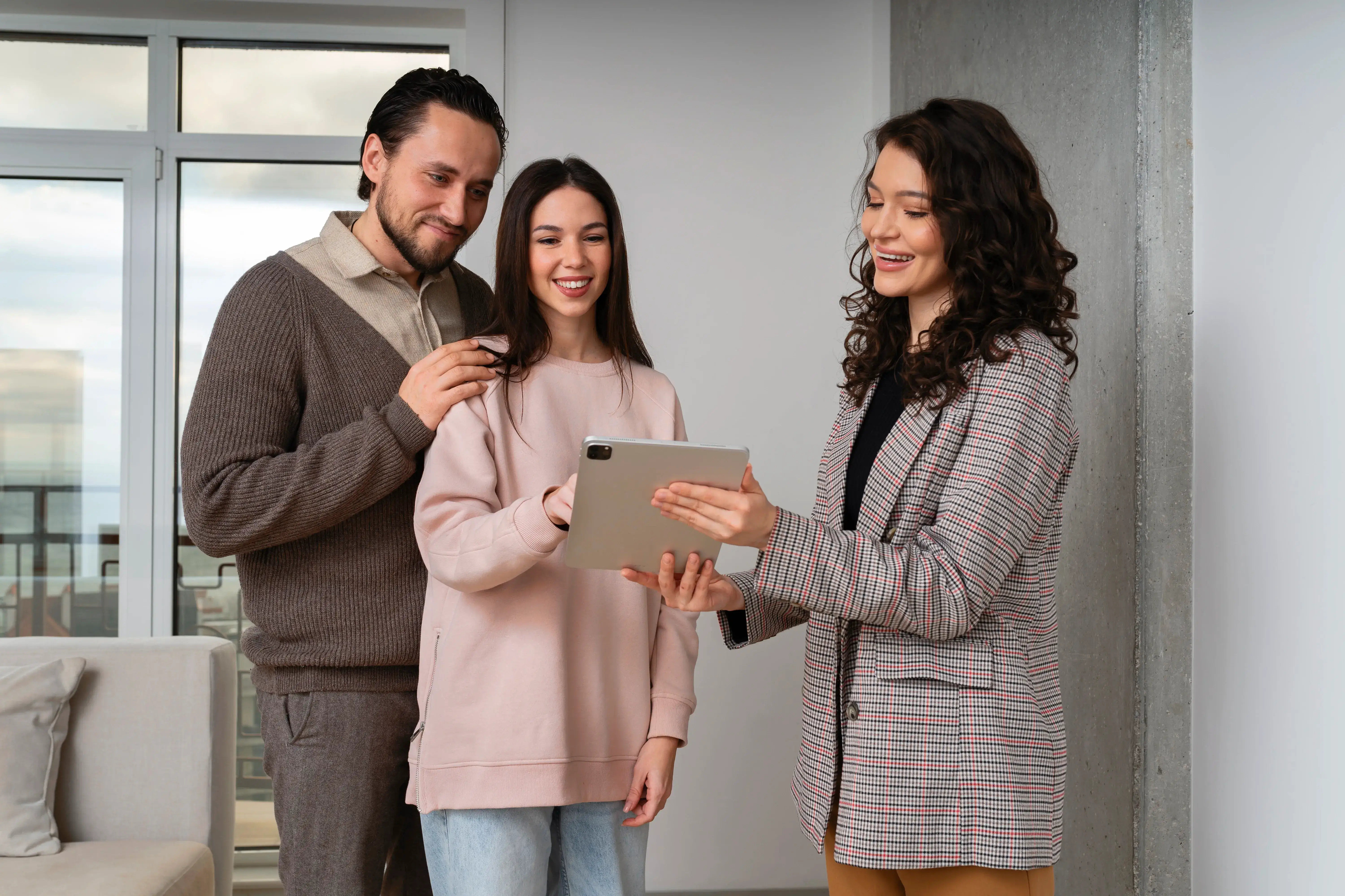A smiling real estate agent gesturing while showing a modern living room to a young couple.