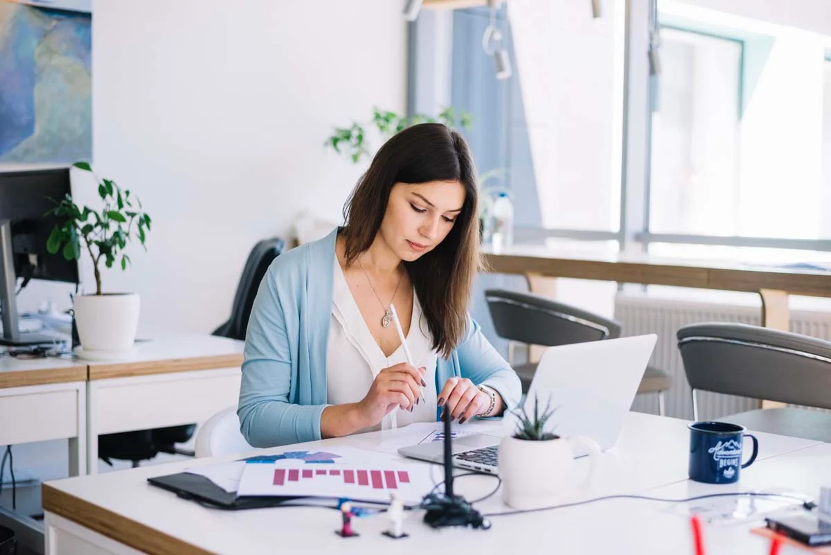 A woman reviews financial charts and graphs on her laptop screen and papers