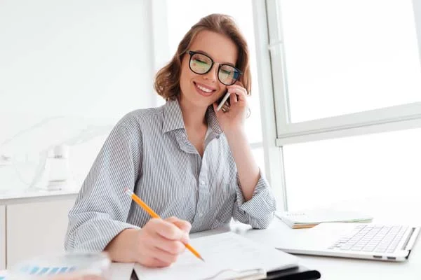 Smiling office worker talking on the phone while taking notes at her desk