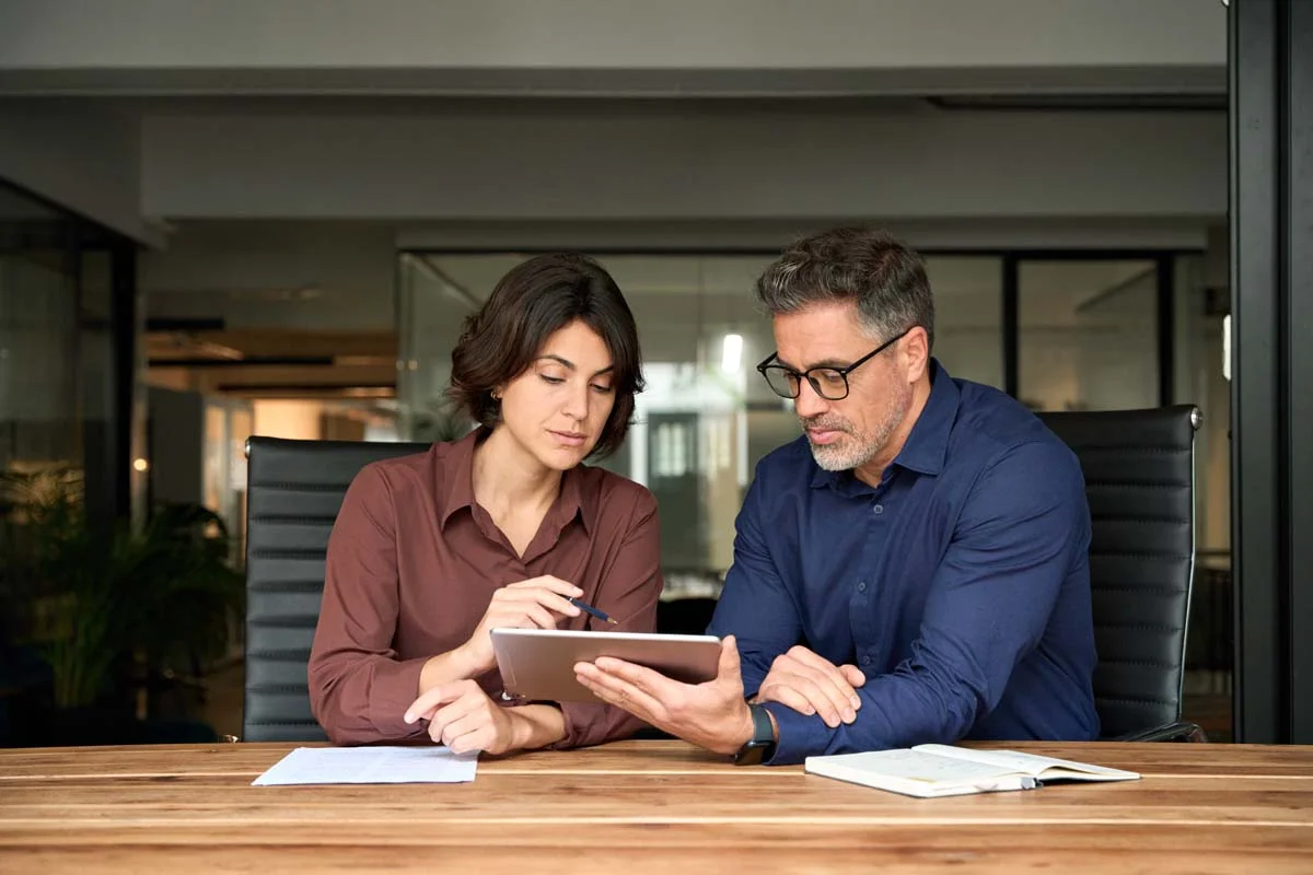 Two business professionals review financial documents together at a desk with a calculator and notebook in the foreground.