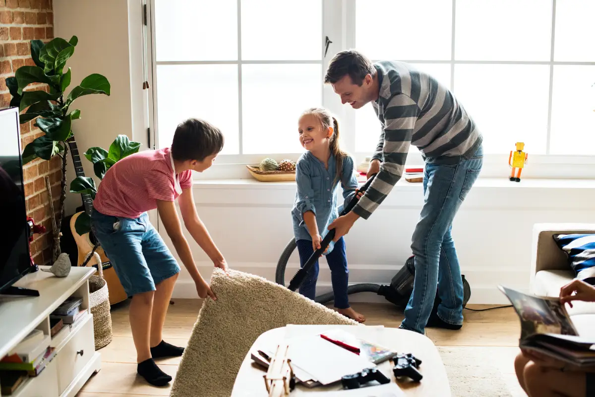 A father and his two children smiling while cleaning the living room together, using a vacuum and lifting a rug.