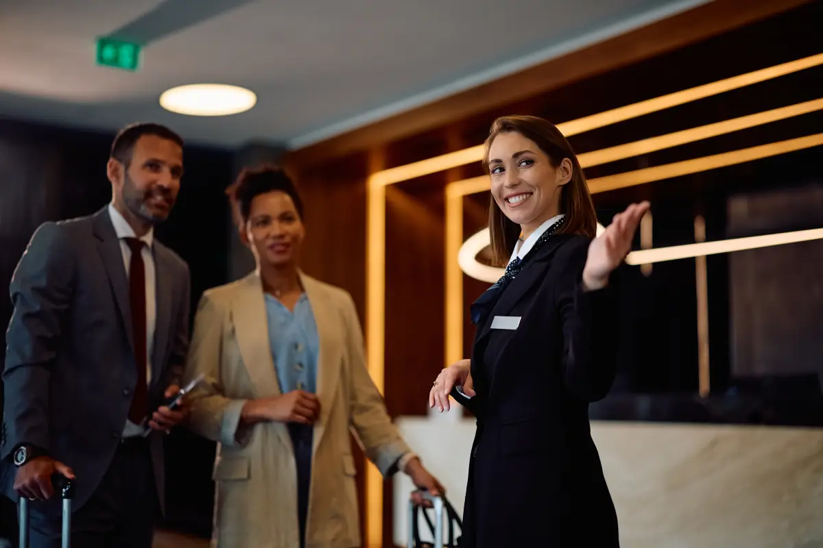 A female hotel concierge or receptionist in a black uniform smiling and gesturing to guide a couple in a bright lobby.