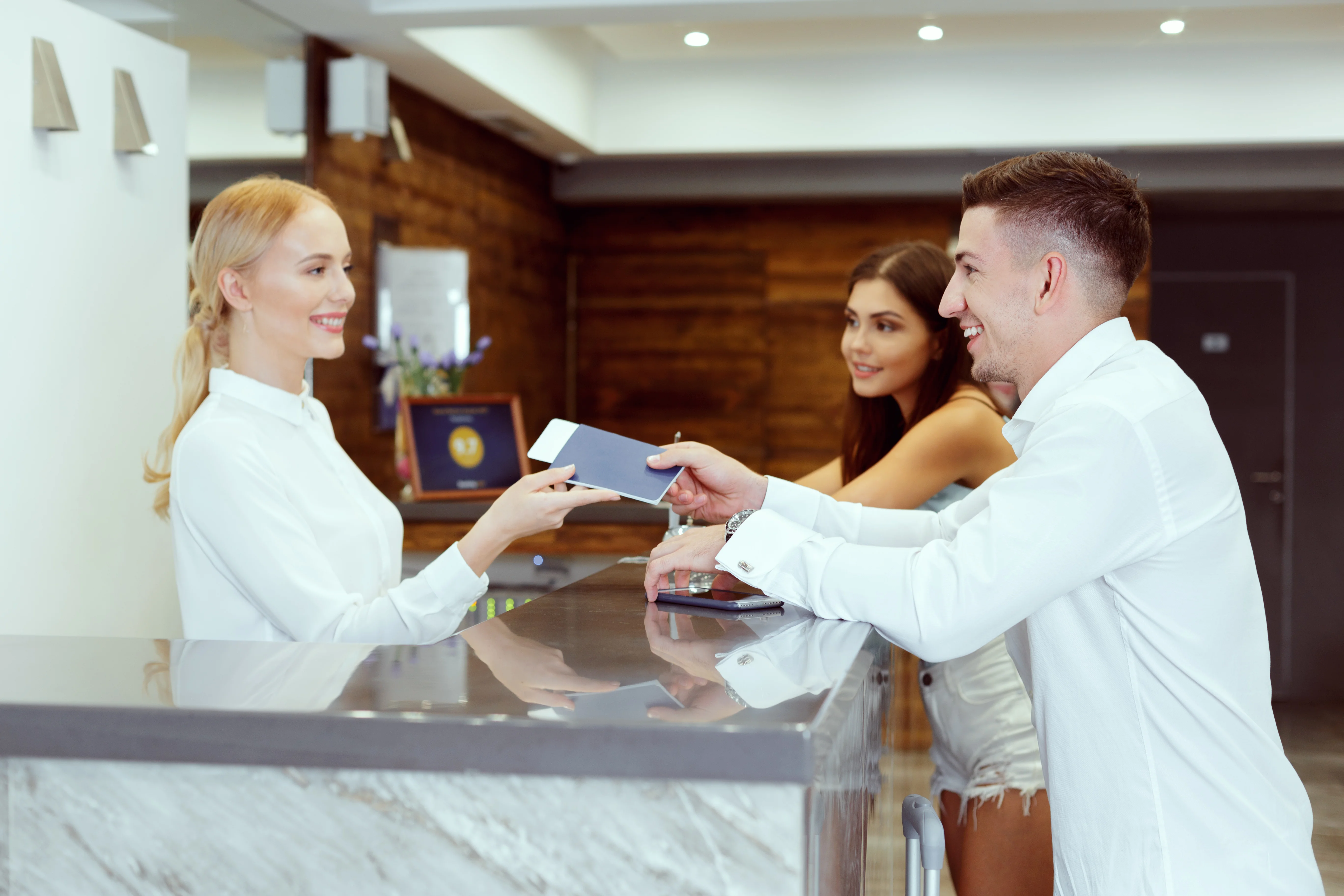 A smiling young couple checking in at a hotel reception desk, with the man receiving a key card from the receptionist.