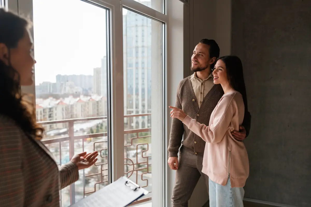 A smiling young couple looks out a large apartment window during a home tour with a real estate agent.