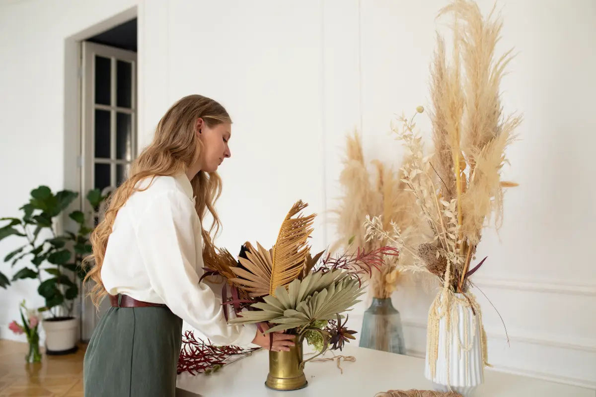 A woman decorates a modern wooden and metal bookshelf, placing dried grass stems into a vase.