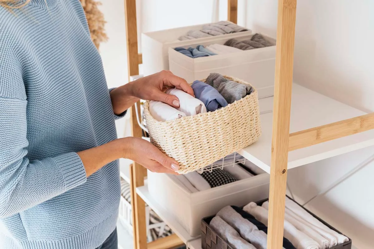 A woman organizes neatly folded blankets, towels, and linens on white wooden shelves using storage baskets