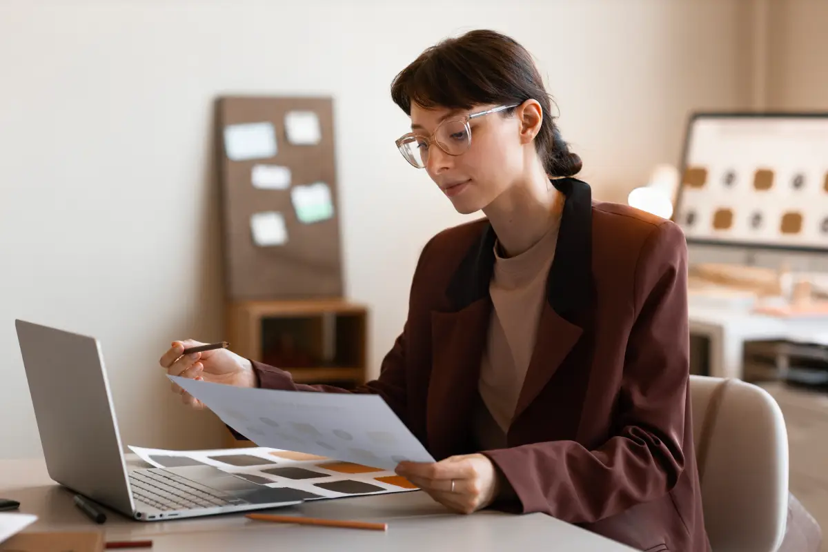 A woman wearing glasses sits at a wooden table, reviewing data on a paper document while comparing it to financial charts open on her laptop screen.