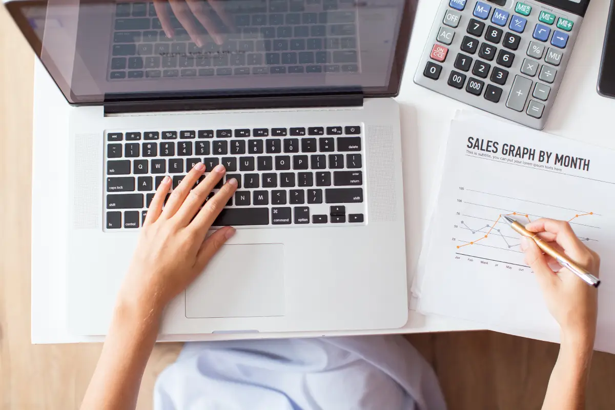 Close-up of a person using a calculator at a wooden desk with financial charts and a laptop.