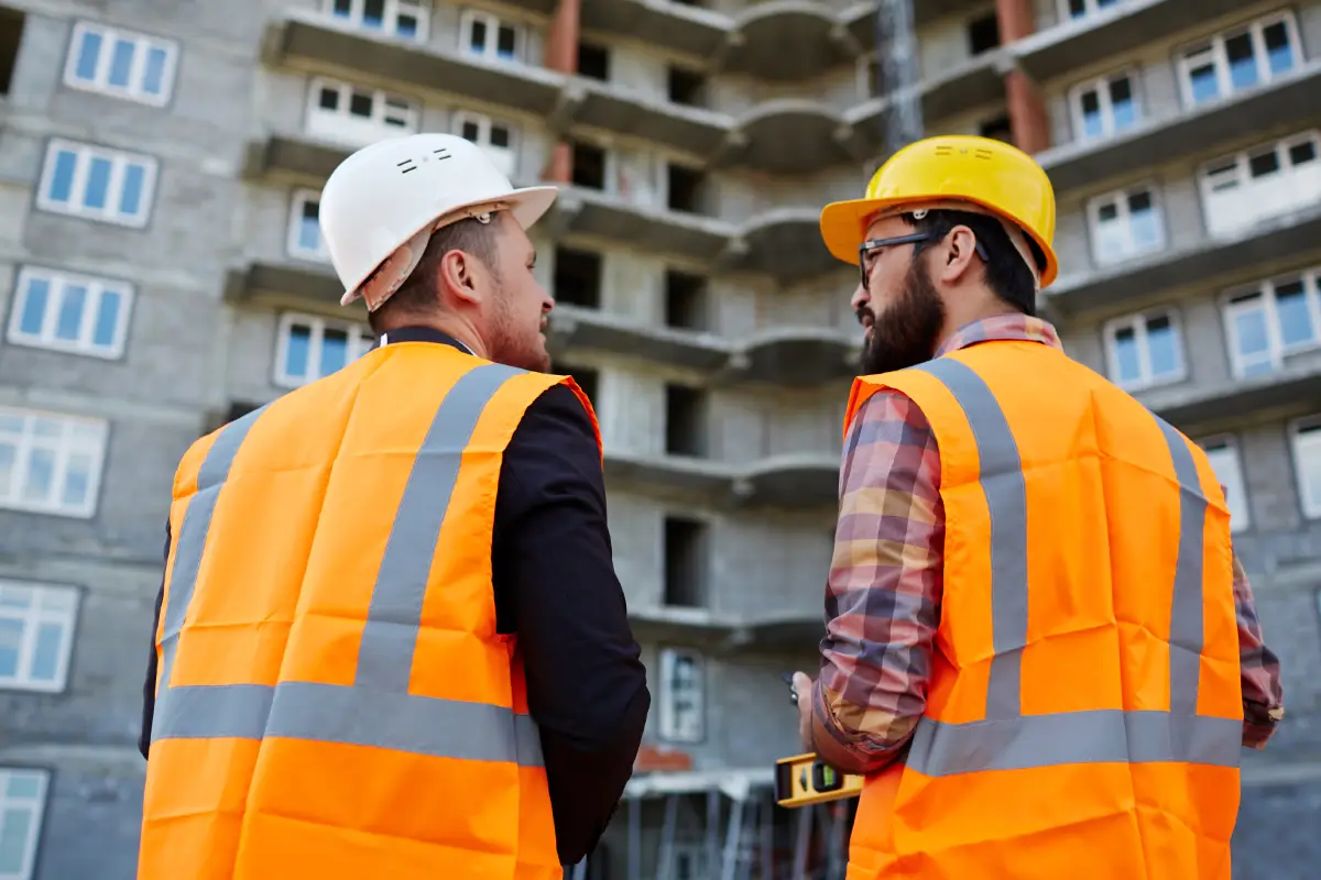 Two male construction professionals wearing yellow hard hats look out a large floor-to-ceiling window in a bright, modern high-rise condo